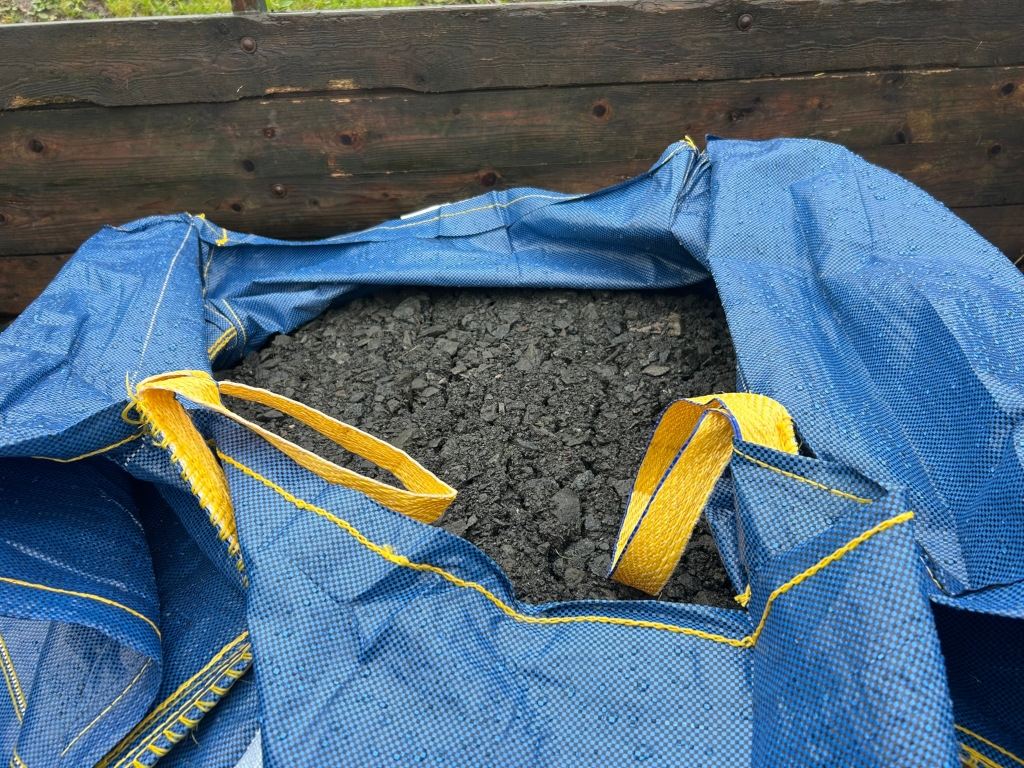 Blue tarp, possibly a large bag or container, filled with dark-coloured, granular material that resembles gravel or crushed stone. The tarp is wet, showing water droplets on its surface, and has yellow straps or handles. The backdrop is a weathered wooden structure.