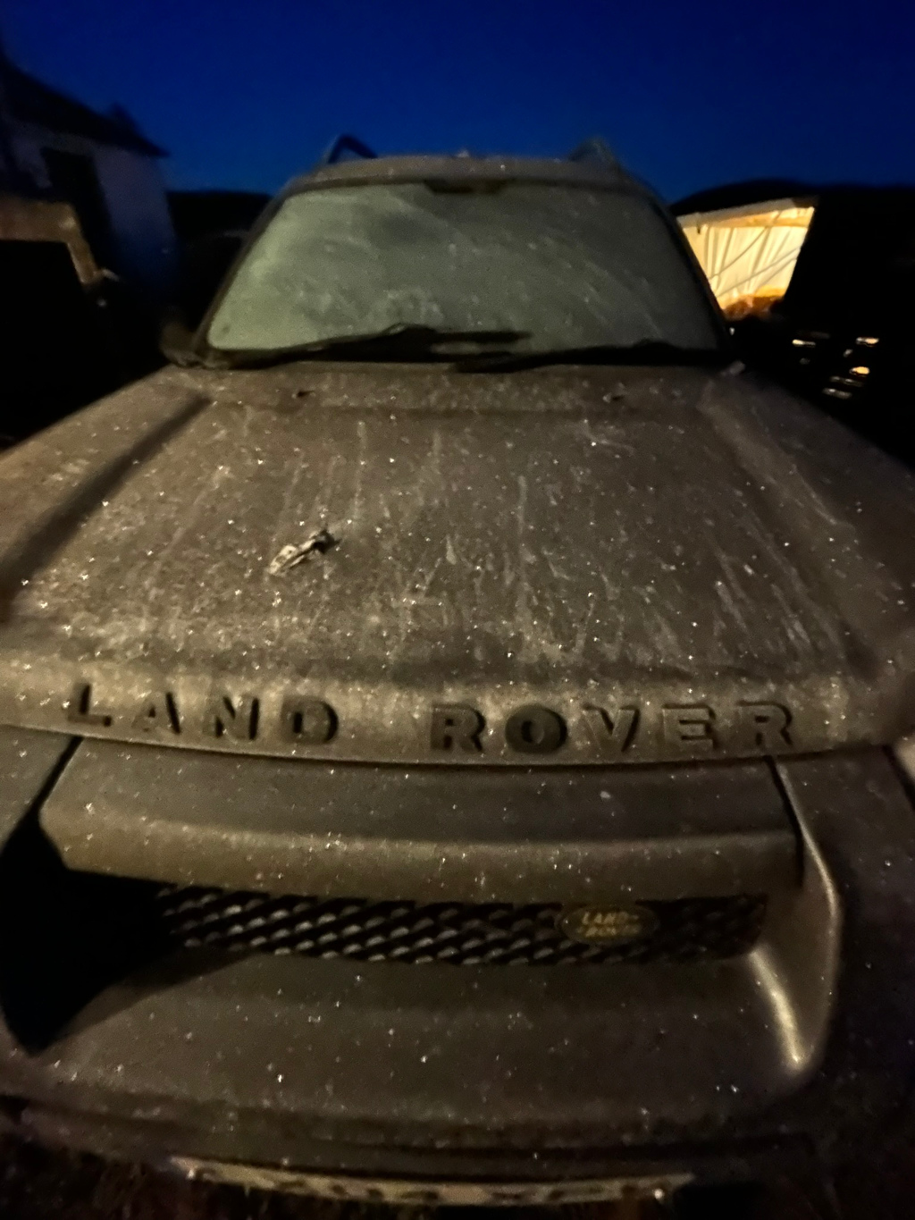 Front of a dark-coloured Land Rover SUV covered in a layer of frost or ice. The frost is visible on the hood, windshield, and grille. The scene is set at night, under a dark sky. A small object, possibly a key, rests on the hood.