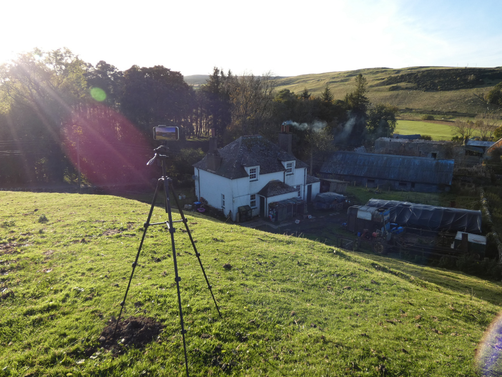 High-angle, long shot of a rural scene. A tripod with a smartphone mounted on top is positioned in the foreground on a grassy hillside. It's pointed towards a white farmhouse and outbuildings nestled in a valley. The background includes rolling green hills, trees, and a hint of late afternoon sun creating lens flare.