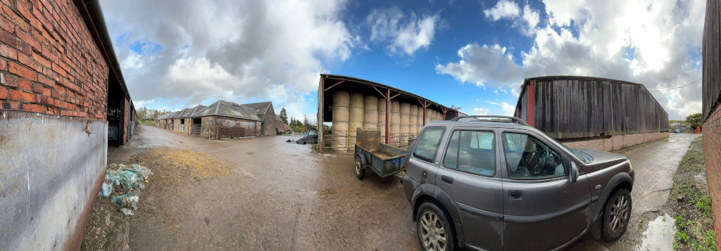 Panoramic view of a farm yard on a partly cloudy day. Several barns of varying ages and construction are visible, one of which contains large bales of hay or straw. A grey Land Rover Discovery is parked in the yard, hitched to a small trailer. The overall impression is of a working farm, possibly in the UK judging by the style of the buildings. The ground is damp, suggesting recent rain. The scene is peaceful and functional, lacking any overt drama or emotional expression.