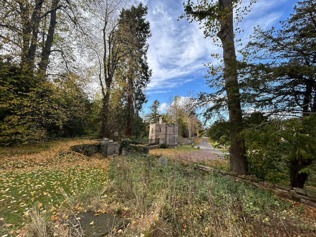 Stone building, possibly a small castle or folly, nestled amongst autumnal trees. Fallen yellow leaves blanket the ground in the foreground, creating a vibrant autumnal scene. A pathway leads towards the building, which is partially obscured by trees and vegetation. The overall mood is peaceful and serene.