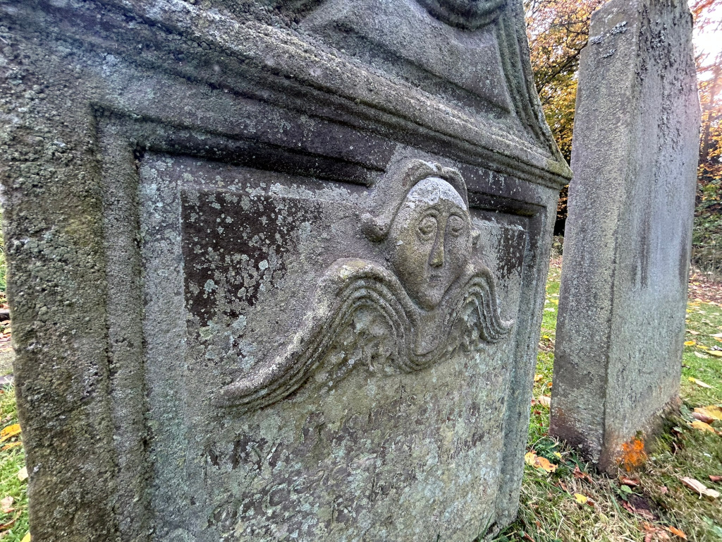 Close-up view of a weathered, moss-covered gravestone. The stone features a carved relief of an angelic or cherubic face with wings, situated within an ornate frame. A second, simpler, upright gravestone is partially visible in the background. The scene is set in a graveyard, with autumn leaves scattered on the ground. The overall impression is one of age, decay, and the passage of time.