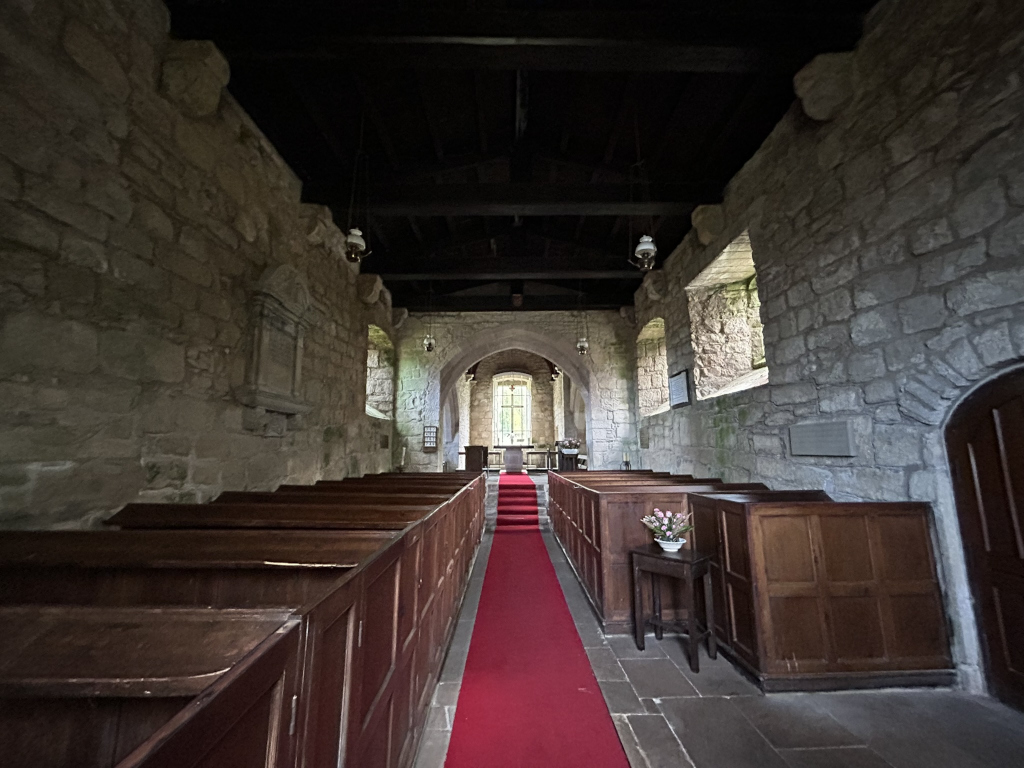 Interior of a stone church. The nave is lined with dark wooden pews, and a red carpet runs down the centre aisle towards the altar area. The church's stone walls are old and uneven, with several small, high windows letting in some light. The overall feeling is one of age, simplicity, and quiet contemplation.