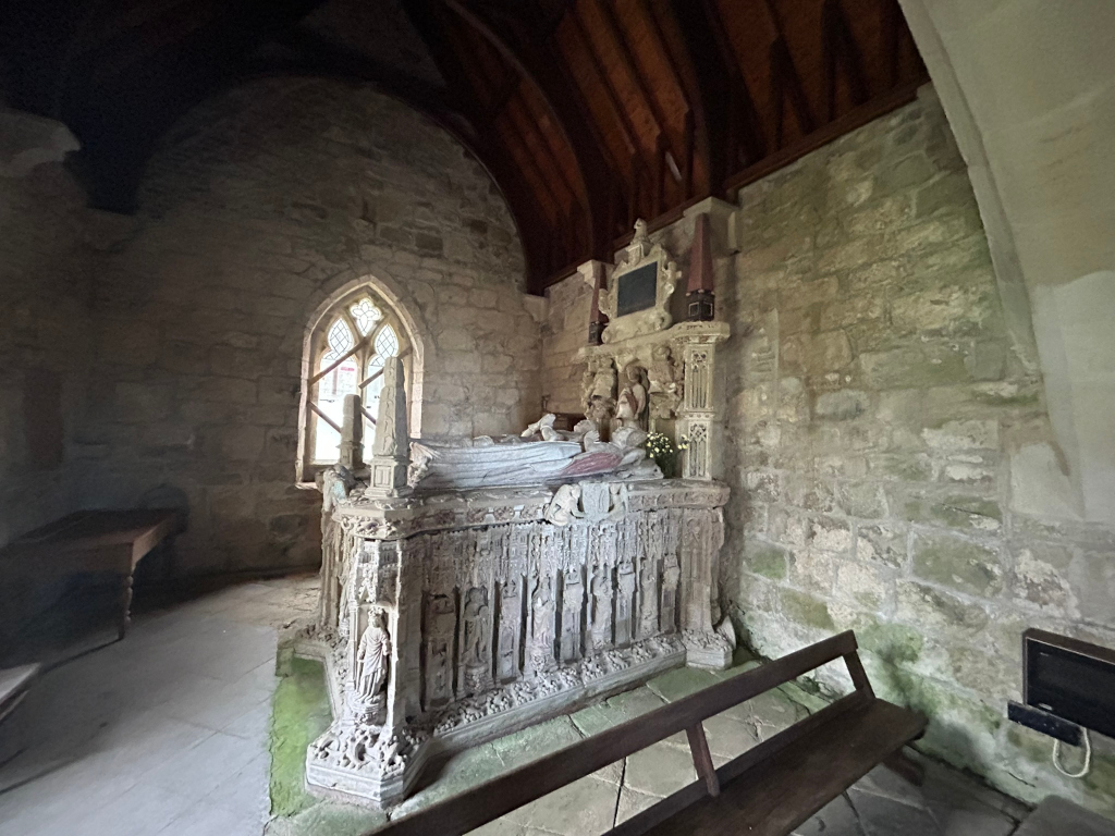 Interior of a stone chapel or church, specifically focusing on an elaborate, ornate tomb. The tomb is large, intricately carved with numerous figures and details, and sits against a rough-hewn stone wall. A small, arched window is visible to the left of the tomb, letting in some light. A simple wooden bench is situated in front of the tomb. The overall atmosphere is one of age, history, and quiet reverence.