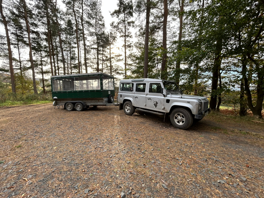Silver Land Rover Defender parked next to a dark-green trailer in a wooded area. The trailer appears to be designed for carrying passengers, possibly for game viewing or similar activities, given the setting and the Land Rover's design. The overall scene suggests a rural or wilderness location, possibly a safari park or nature reserve.