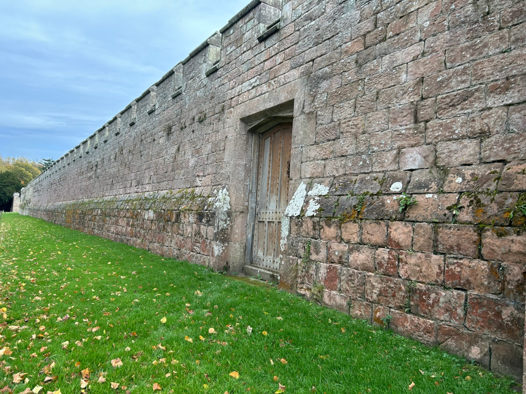 Long, high stone wall, possibly part of a castle or other historical structure. The wall is made of reddish-brown stone blocks and shows signs of age and weathering, including moss in places.  Set into the wall is a single, wooden door, appearing old and sturdy.  The foreground shows a grassy area with scattered fallen leaves, suggesting autumn. The overall impression is one of age, history, and perhaps a sense of seclusion or mystery.