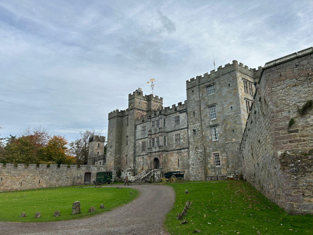 Large, imposing stone castle on an overcast day. The castle is multi-storied with crenellated walls and numerous windows.  A dark green vintage-looking vehicle and a horse-drawn cart are parked in the courtyard. A person can be seen walking near the vehicles. The castle is surrounded by a high stone wall and grassy areas. The overall impression is one of history and grandeur.