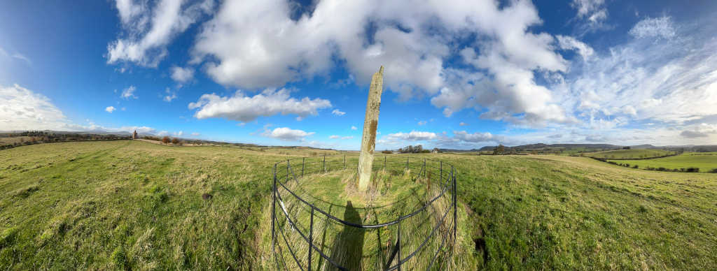 Panoramic view of a grassy field under a bright, partly cloudy sky. A tall, slender stone, possibly a standing stone or monolith, is the central focus, enclosed by a simple black metal fence. The surrounding landscape is gently rolling hills, indicative of a rural or agricultural setting. The overall impression is one of tranquillity and openness, with the stone suggesting a historical or archaeological significance to the location