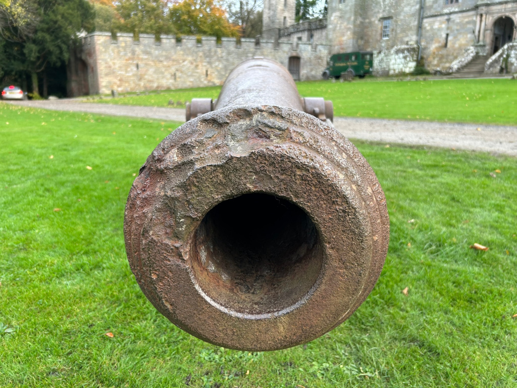 Close-up view of the muzzle of an old, rusty cannon. The cannon is situated on a grassy lawn in front of a large stone castle or fortress. The background is slightly out of focus, drawing attention to the texture and detail of the cannon's weathered surface. A vehicle is visible in the far background, adding a sense of scale and context.