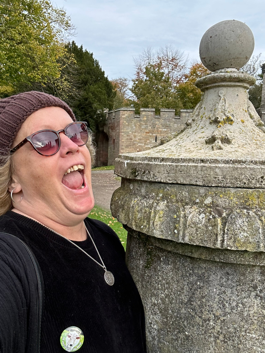 Leonie in a brown knit hat and sunglasses, her mouth wide open in a joyful expression. She is standing next to a weathered stone pillar or post, which appears to be part of a larger stone structure (possibly a wall or fence) visible in the background. The background also shows some trees and what appears to be an old stone castle or fortified building which is partially visible. She is wearing a black top and a silver necklace. There is also a small circular button or badge visible on her sweater. The overall impression is one of a happy, spontaneous moment captured outdoors in a historical setting.