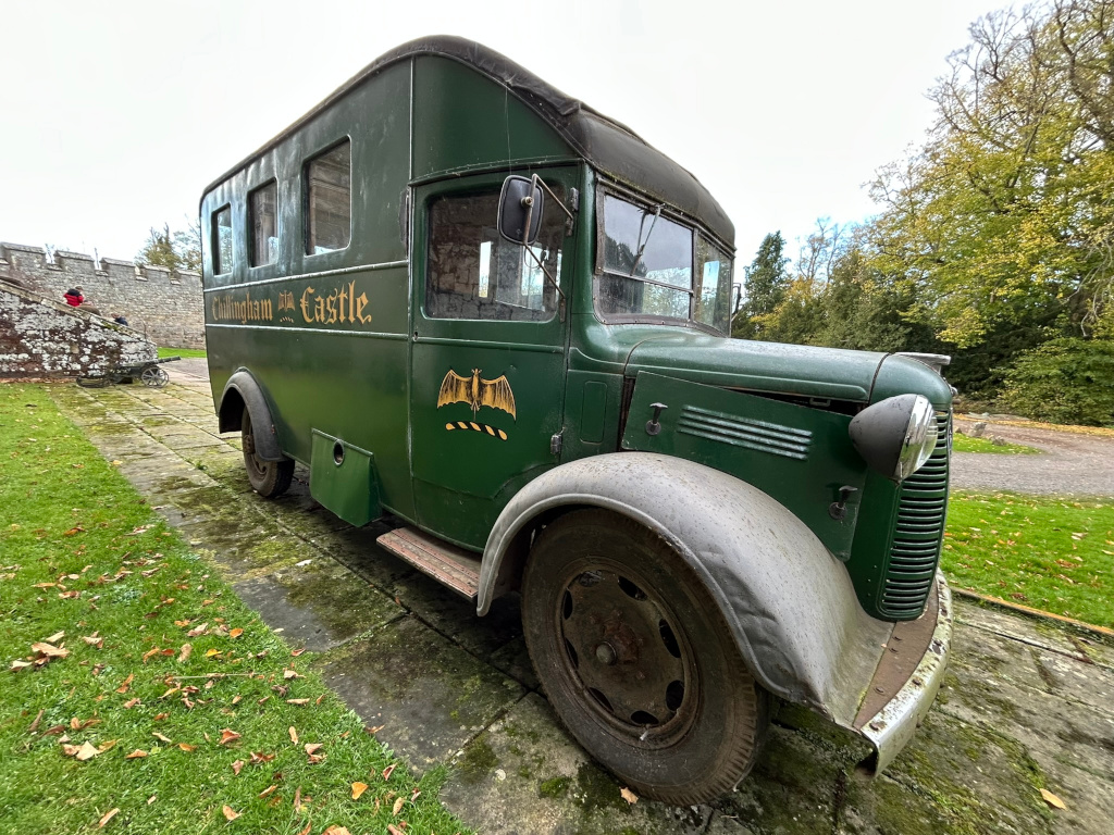 Vintage, dark green van parked on a paved area next to a stone wall. The van is labelled Chillingham Castle in gold lettering and has a bat insignia. The setting appears to be the grounds of a castle, with autumnal foliage and a glimpse of a person in the background. The overall impression is one of age, history, and possibly a connection to a theme of Gothic or mystery, given the castle setting and bat symbol.