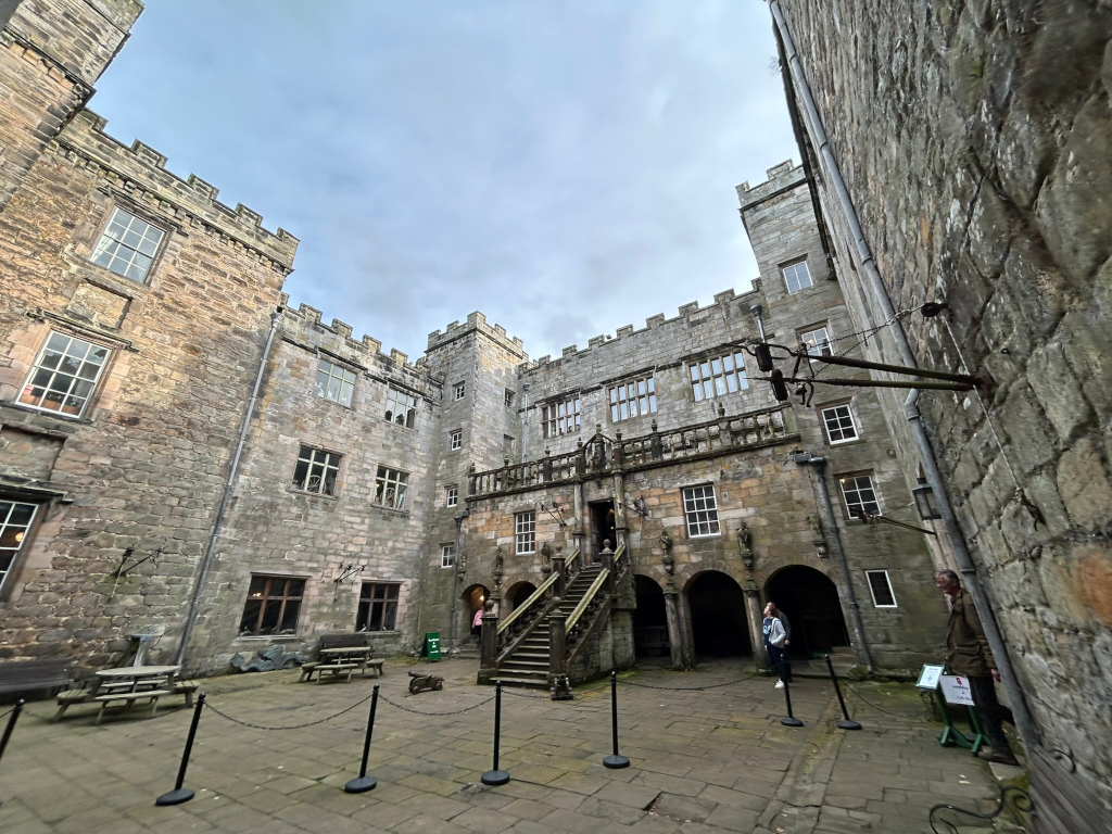 Interior courtyard of a stone castle. The courtyard is paved, and there are stone walls, stairs leading to an upper level, and several arched passageways. Two people are visible in the courtyard, and there's some simple outdoor furniture. The overall style is medieval or historic.
