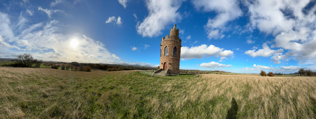Panoramic view of a landscape dominated by a stone tower, reminiscent of a folly or small castle. The tower stands in a field of tall, dry grass under a bright, sunny sky with scattered fluffy white clouds. The background shows a gently rolling, grassy landscape extending to a distant horizon.