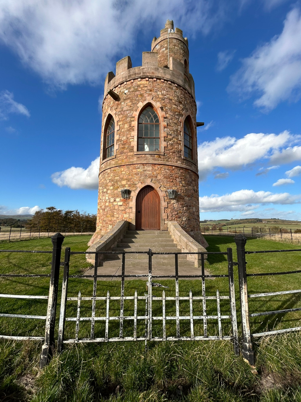 Stone tower, reminiscent of a small castle or folly, standing in a grassy field under a partly cloudy blue sky. The tower is circular with crenellations at the top, arched windows, and a wooden door. A weathered metal fence with a gate stands in the foreground. The scene is peaceful and evokes a sense of quiet history or solitude.