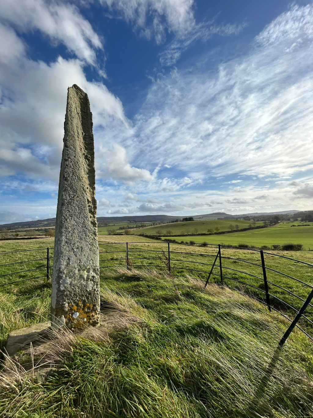 Tall, slender, weathered stone standing upright in a grassy field, enclosed by a black metal fence. The sky is a brilliant blue with fluffy white clouds. Rolling green hills stretch out into the distance under the expansive sky. The overall impression is one of serenity and the quiet majesty of the landscape. The stone itself appears ancient, possibly a standing stone or menhir, suggesting a historical or possibly even mystical significance.
