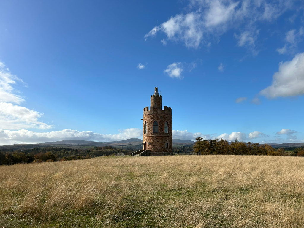 Solitary, stone tower, reminiscent of a small castle or folly, standing in a field of tall, dry, golden grass under a bright blue sky with scattered fluffy white clouds. In the distance, rolling hills are visible under the clear sky. The overall impression is one of serenity and tranquillity, accentuated by the contrast of the man-made structure against the expansive natural landscape.