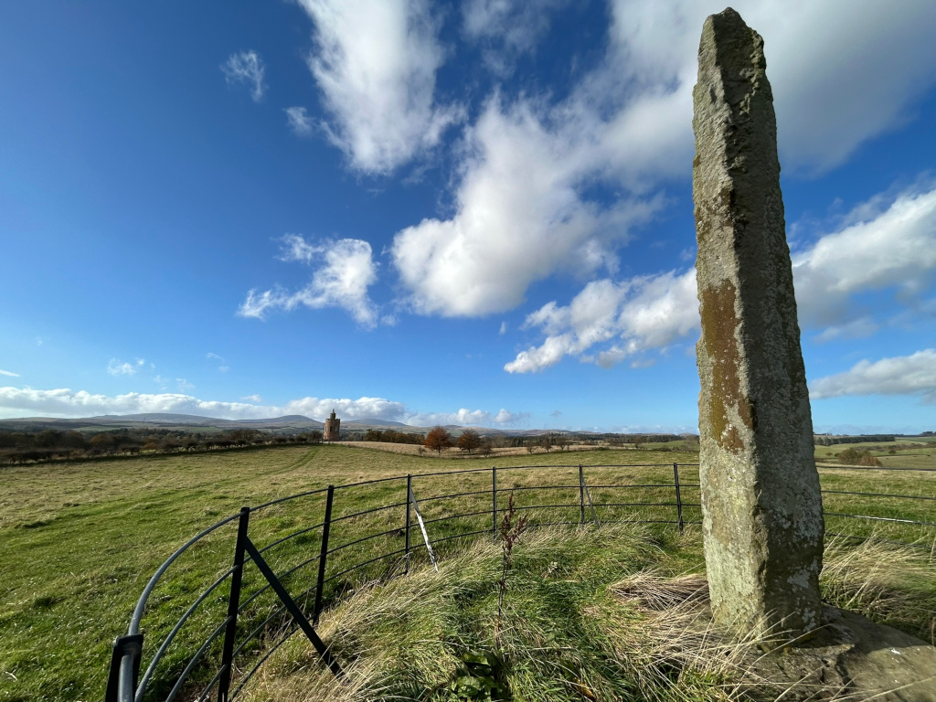 Tall, slender standing stone in a grassy field, enclosed by a black metal fence. In the far distance, a small, brick tower-like structure is visible against a backdrop of rolling hills under a bright blue sky with fluffy white clouds. The scene evokes a sense of history and peaceful rural landscape.