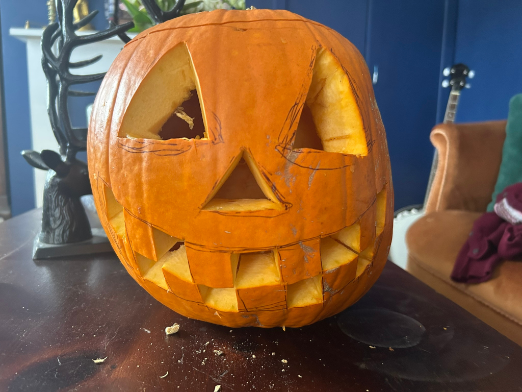 Carved Halloween pumpkin sitting on a dark brown wooden table. The pumpkin's design is a grinning face, with the eyes, nose, and mouth only partially cut out; the lines indicating the cuts are still visible. The background is slightly blurred but shows a dark blue wall, part of a deer-head ornament, and a portion of an armchair. The overall impression is one of a work in progress, a pumpkin being prepared for Halloween.