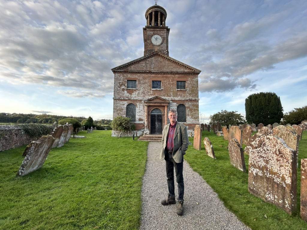 Charlie standing in front of a brick church in a graveyard. The church is old, with a clock tower, and the gravestones are weathered. He is dressed in casual attire and appears to be looking directly at the camera. The setting suggests a historical or possibly sombre mood. The overall composition is balanced and well-lit.