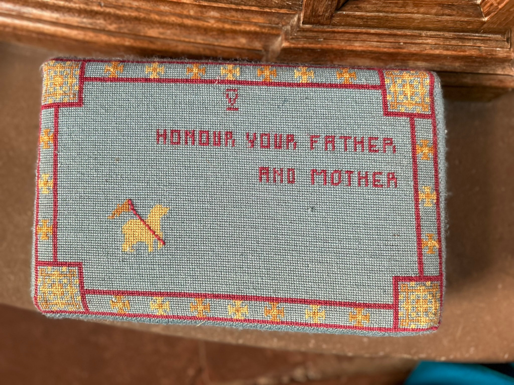 Needlepoint cushion or kneeler. It's rectangular and features a muted blue-green background with the phrase Honour your Father and Mother embroidered in red. A small, yellow embroidered lamb with a flag or banner is also present. The border is decorated with a geometric pattern in red and gold thread. The overall style suggests a religious or traditional context.