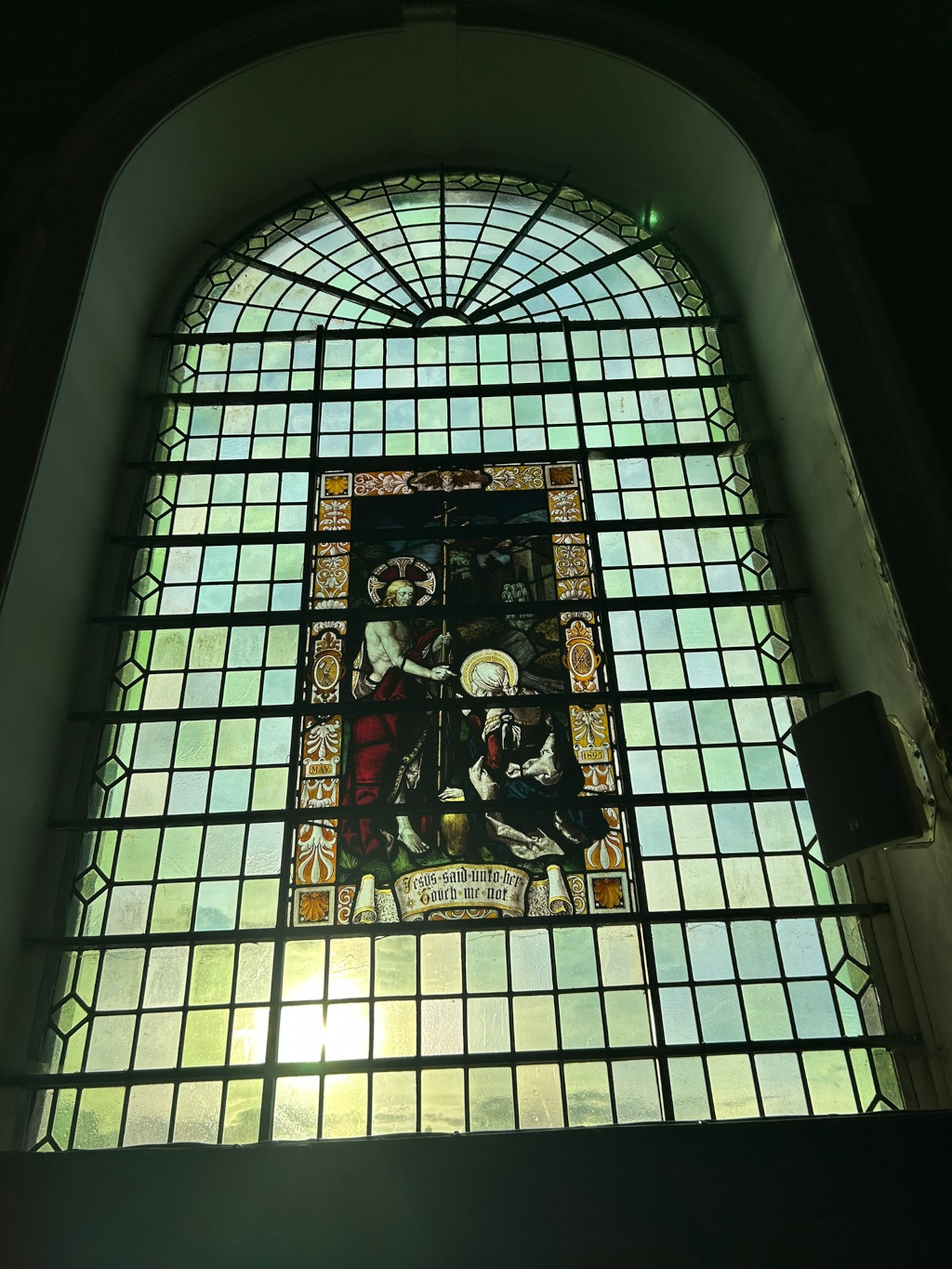 Stained-glass window, arched at the top, set within a larger, darker arched window frame. The stained glass depicts a scene from the Bible, likely the Resurrection, with Jesus appearing to Mary Magdalene. The text at the bottom reads Jesus said unto her Touch me not. The overall style is traditional, suggesting a church or religious building. Sunlight streams through the window from behind, adding to the contemplative atmosphere.