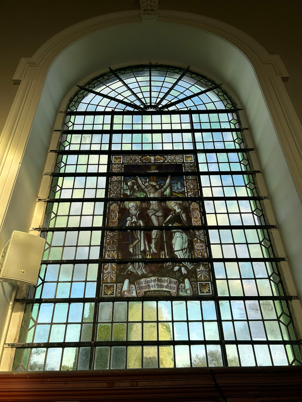Large arched stained-glass window, typical of a church or cathedral. The central panel depicts a scene of the crucifixion of Jesus Christ, with Mary and John flanking him. Below the depiction is an inscription referencing the cleansing power of Jesus' blood. The window is divided into many smaller panes by a dark metal framework, and the overall style suggests a traditional, possibly historical, setting. A modern speaker is visible mounted on the wall to the left of the window.