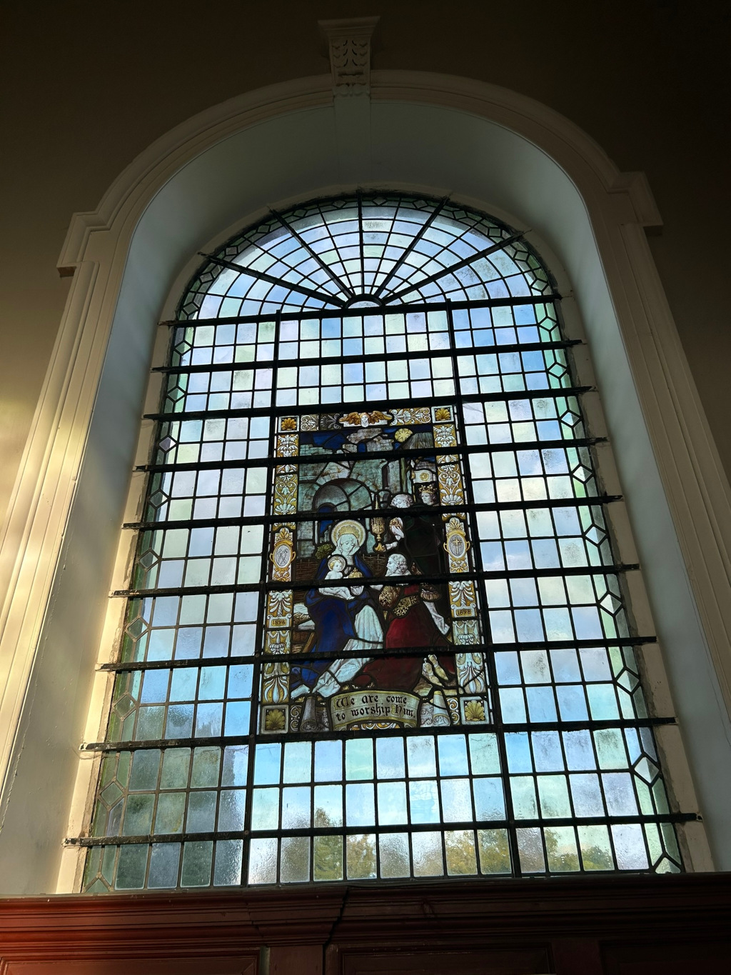 Large arched stained-glass window, characteristic of a church or other religious building. The main feature is a central panel depicting the Adoration of the Magi, a biblical scene showing the three wise men presenting gifts to the baby Jesus. The panel is surrounded by smaller, clear glass panes creating a grid pattern within the arched frame. At the bottom of the central panel, an inscription reads We are come to worship Him. The overall style suggests a traditional, possibly Victorian-era, aesthetic. The image is well-lit, highlighting the colours and detail of the stained glass.