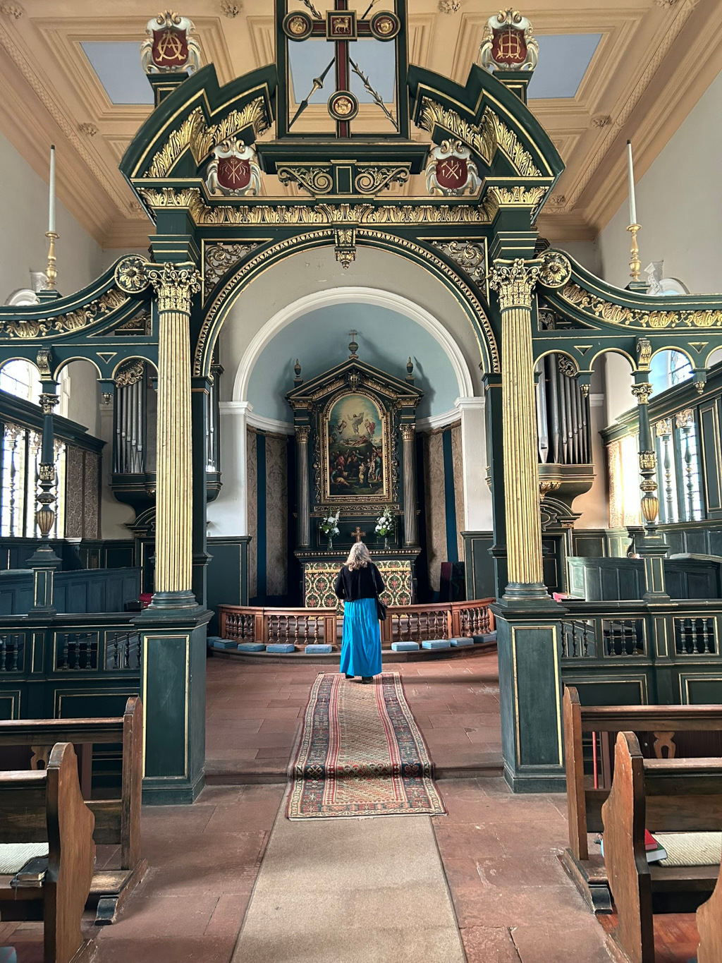 Interior of a church, specifically focusing on the nave and altar area. Leonie in a long blue skirt and dark jacket stands with her back to the camera, facing the altar. The church features elaborate, dark green and gold woodwork, with a richly decorated reredos behind the altar containing a painting depicting a religious scene. A rug stretches from Leonie's position towards the altar. The overall impression is one of quiet contemplation and reverence within a historic and beautifully crafted religious space.