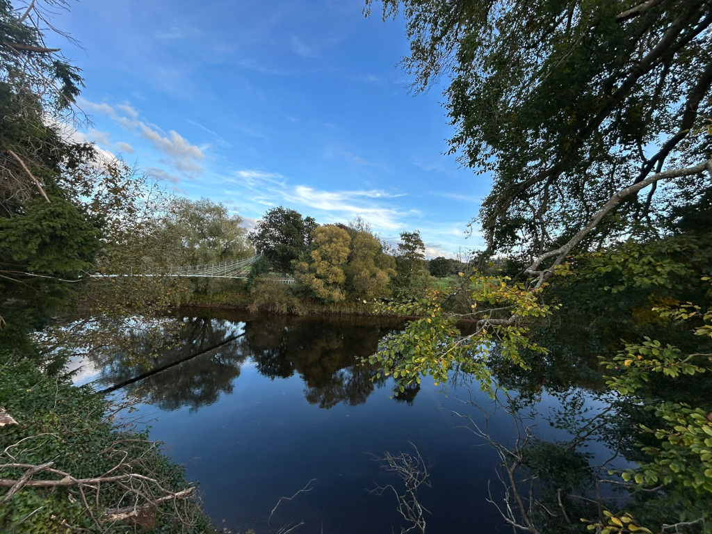 Tranquil scene of a river reflecting the surrounding trees and a suspension bridge in the background under a mostly clear blue sky. The water is dark and calm, mirroring the autumnal colours of the trees. The overall mood is peaceful and serene. The viewpoint is from the riverbank, partially obscured by overhanging branches.