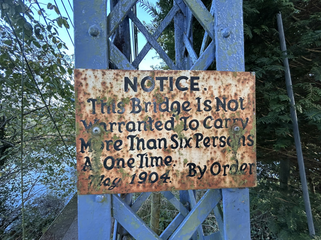 Rusty, aged metal sign affixed to a blue metal bridge structure. The sign, worn by time and the elements, displays a notice that reads: NOTICE. This Bridge is Not Warranted to Carry More Than Six Persons At One Time. May 1904. By Order. The photo includes the surrounding natural environment of trees and what seems to be a river or water body visible in the background, adding to the sense of age and history that the image conveys. The overall effect is one of rustic charm and historical significance. The rusty sign itself speaks to the passage of time, adding a layer of visual interest to the scene.