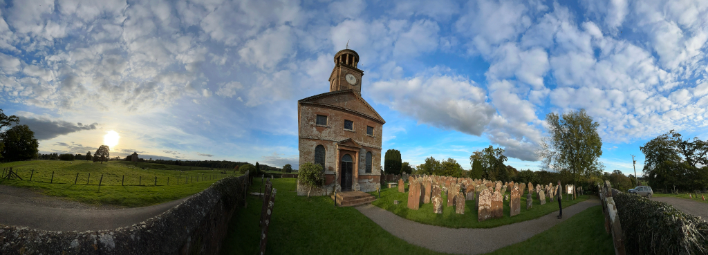 Panoramic view of a brick church with a clock tower. The church is situated in a green field, surrounded by a graveyard with numerous weathered headstones. The sky is mostly blue with a scattering of fluffy white clouds. The overall scene is peaceful and evokes a sense of quiet countryside serenity. The sunlight suggests it is likely late afternoon or early evening.