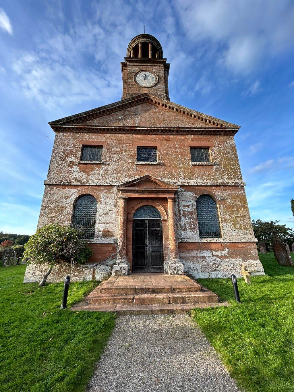 Sandstone church, possibly in the UK, with a clock tower. The church is aged, showing weathering and lichen. A graveyard is visible in the foreground and background. The overall aesthetic is one of quiet, historical dignity.