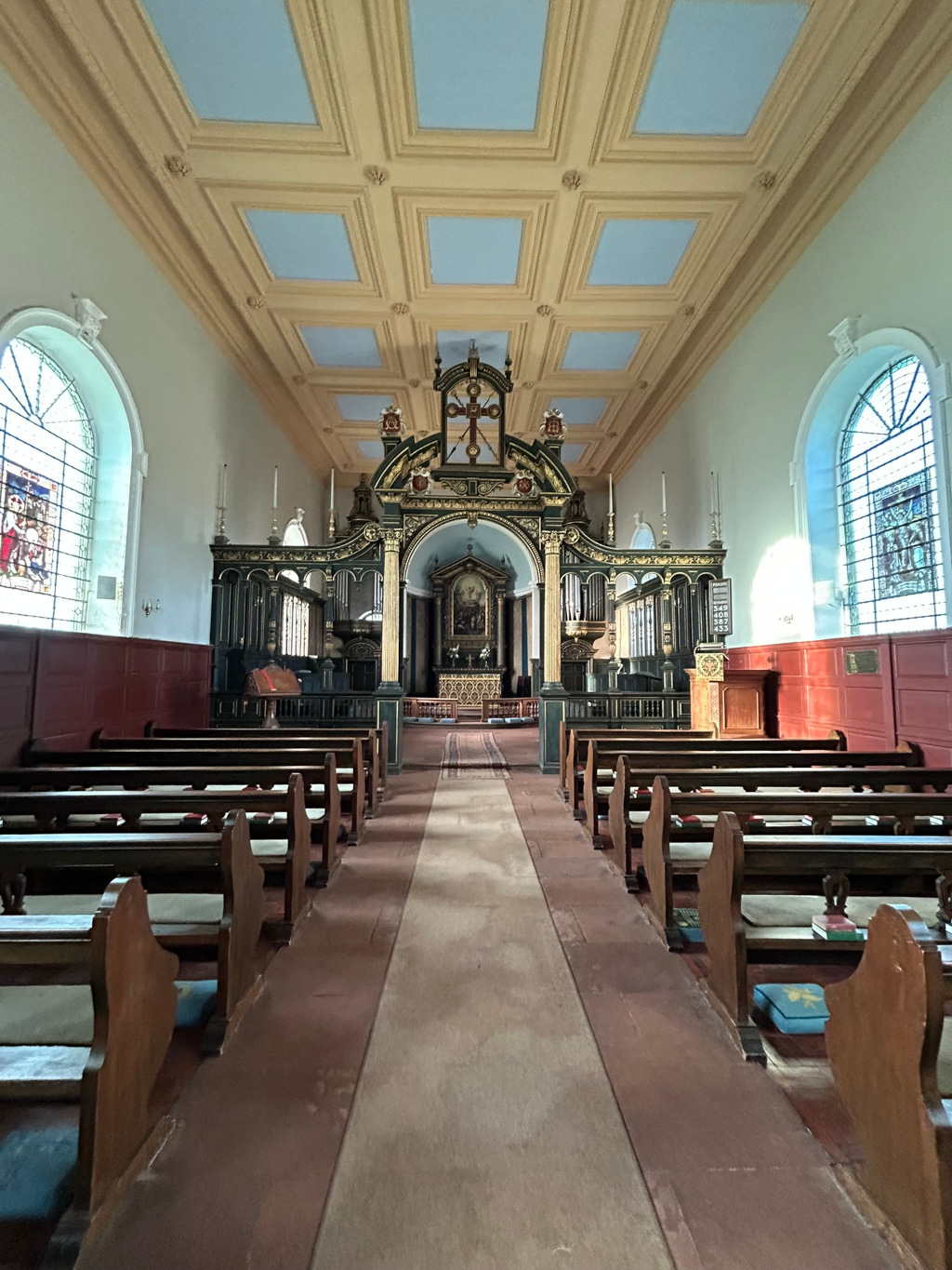 Interior of a church. The perspective is from the nave, looking towards the altar.  The church features an ornate, dark wood reredos (the decorative screen behind the altar) and rows of simple wooden pews. The ceiling is coiffured with a light beige and blue pattern. Stained-glass windows are visible on either side of the nave. The overall impression is one of quiet dignity and historical significance.