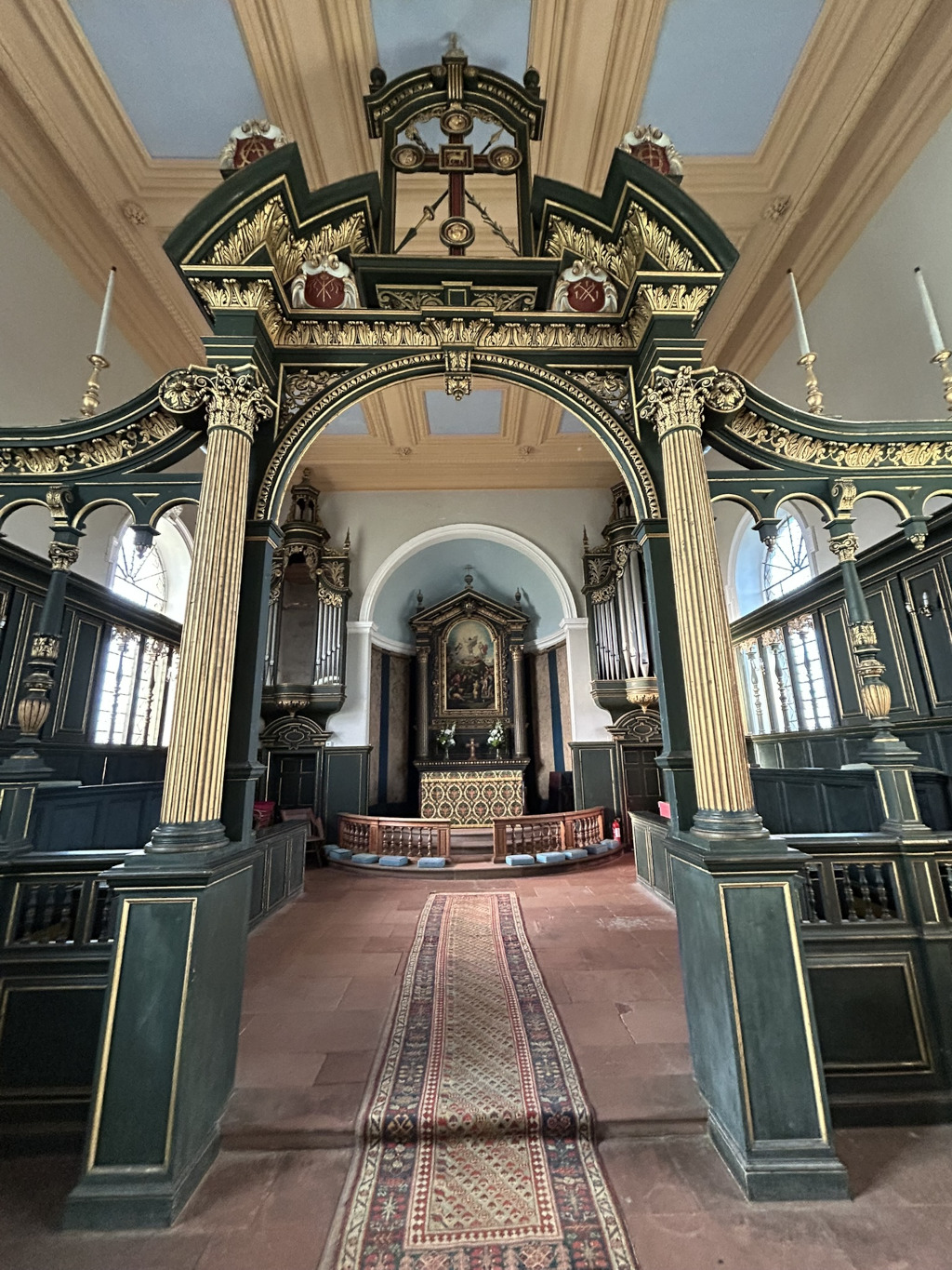Interior of a church, specifically the chancel area. The scene is dominated by a large, ornate, dark green and gold structure, possibly a screen or pulpit, which frames the altar and the main sanctuary. A runner carpet leads from the viewer's perspective towards the altar. The overall style suggests a historical or classical architectural design. The altar itself is relatively simple compared to the elaborate surrounding structure.