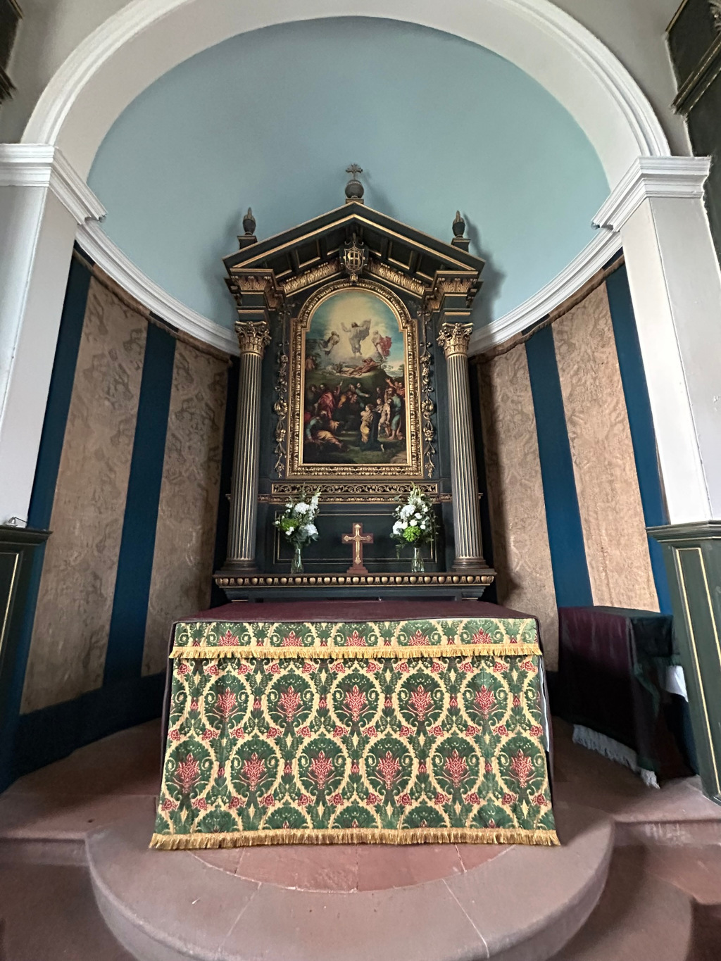 Altar in a chapel or church. The altar is adorned with an ornate, dark-wood reredos featuring a painting depicting a biblical scene, possibly a depiction of the ascension of Christ or a similar religious event.  The altar cloth is a richly patterned fabric with green and gold tones. The room's walls are a dark teal with vertically striped, aged tapestry or wall covering. The overall atmosphere is one of quiet reverence and historical significance.