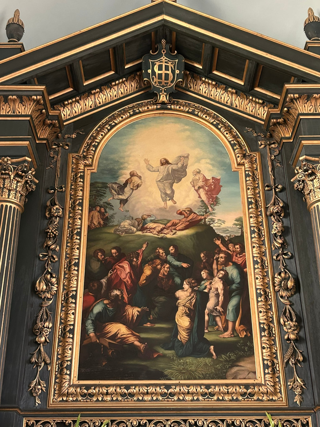 Painting of the Ascension of Jesus Christ, set within an ornate, dark-green and gold frame. Jesus is depicted ascending into heaven, surrounded by clouds, while a group of disciples looks on from below in a state of awe and wonder. The artistic style appears to be reminiscent of Renaissance painting. The frame itself is a significant element, showing intricate carvings and gilding, suggesting a place of religious importance, likely within a church or chapel.