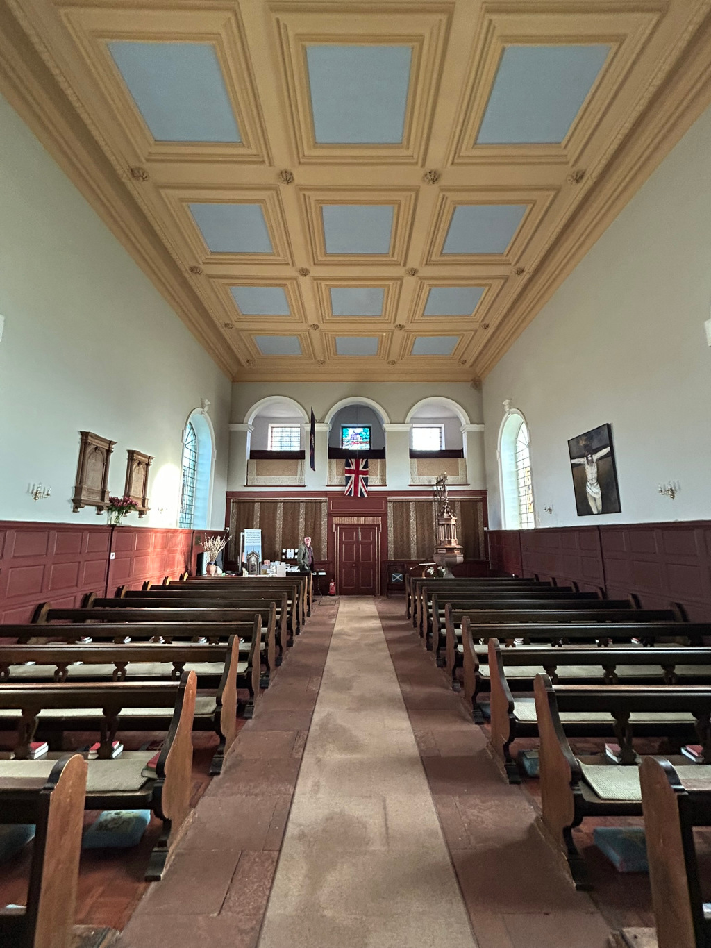 Interior of a church. The view is from the aisle, looking toward the altar. The church features dark wooden pews arranged in rows, a beige and blue coffered ceiling, and walls painted a light grayish-beige. A Union Jack flag hangs on the wall behind the altar. There's a small table with items on it near the center of the aisle, and a person standing near it. The walls have some wood paneling and artwork (including a depiction of the crucifixion of Jesus) hangs on the walls. The overall atmosphere is calm and quiet.