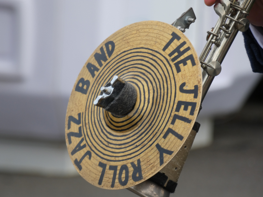 Close-up of a cymbal belonging to The Jelly Roll Jazz Band. The cymbal is gold-coloured with black lettering indicating the band's name. A portion of a musical instrument, possibly a clarinet or saxophone, is visible in the background, suggesting the cymbal is part of a musical setup.
