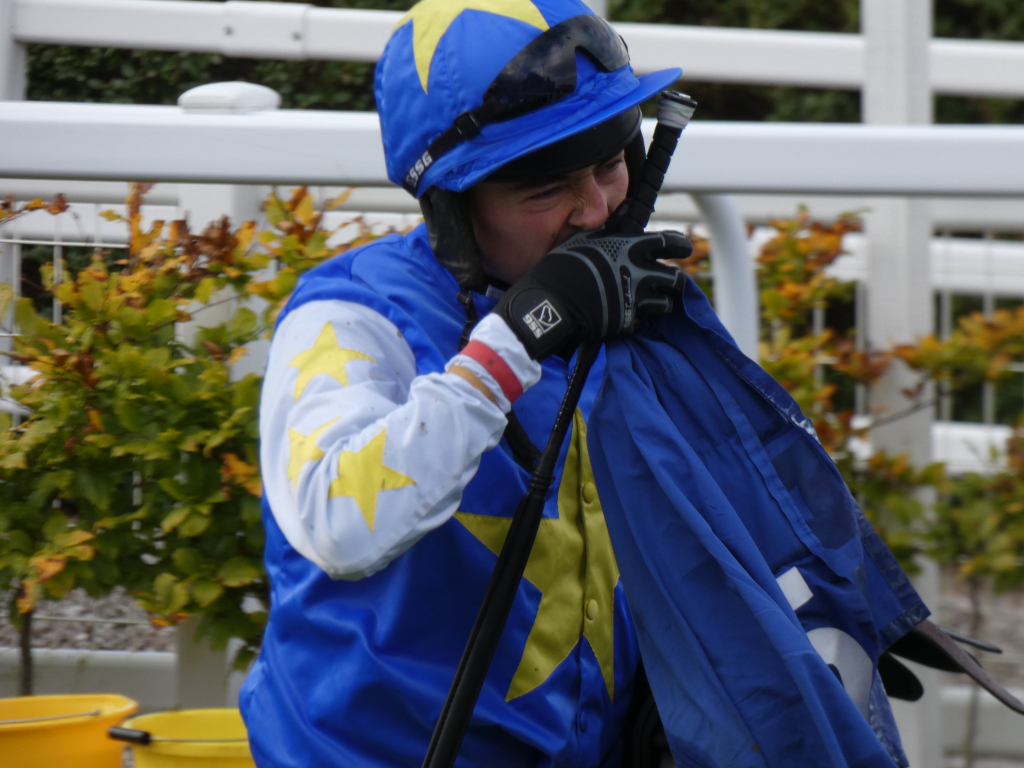 Jockey, wearing a blue and yellow racing silks, holding a blue silks jacket over their shoulder. They are wearing a helmet and gloves. Their face is partially obscured, and they appear to be wiping their nose or mouth. The background suggests a racecourse setting. The overall mood is one of post-race weariness or disappointment.