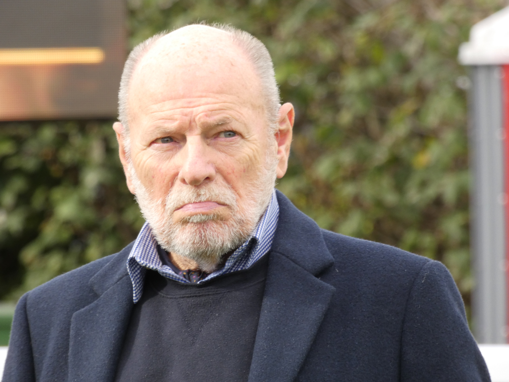 Close-up of an older, bald Caucasian man with a serious expression. He has a gray beard and is wearing a dark navy blazer over a dark gray sweater and a light blue and white patterned shirt. The background is blurred, showing out-of-focus greenery and what appears to be a portion of a scoreboard or electronic display. The man's gaze is direct, and his expression conveys a sense of contemplation or perhaps slight displeasure.