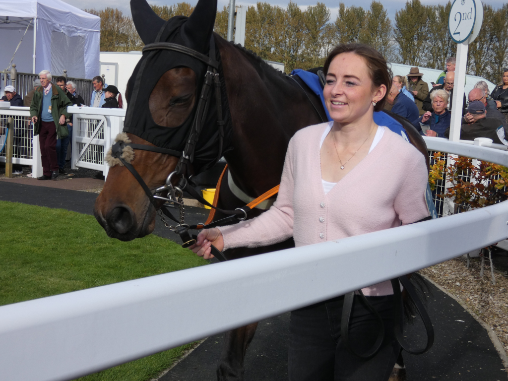 Woman in a pink cardigan leading a brown horse with a black hood. They are at a racetrack, with spectators visible in the background. The woman appears to be a jockey or someone involved in horse racing.