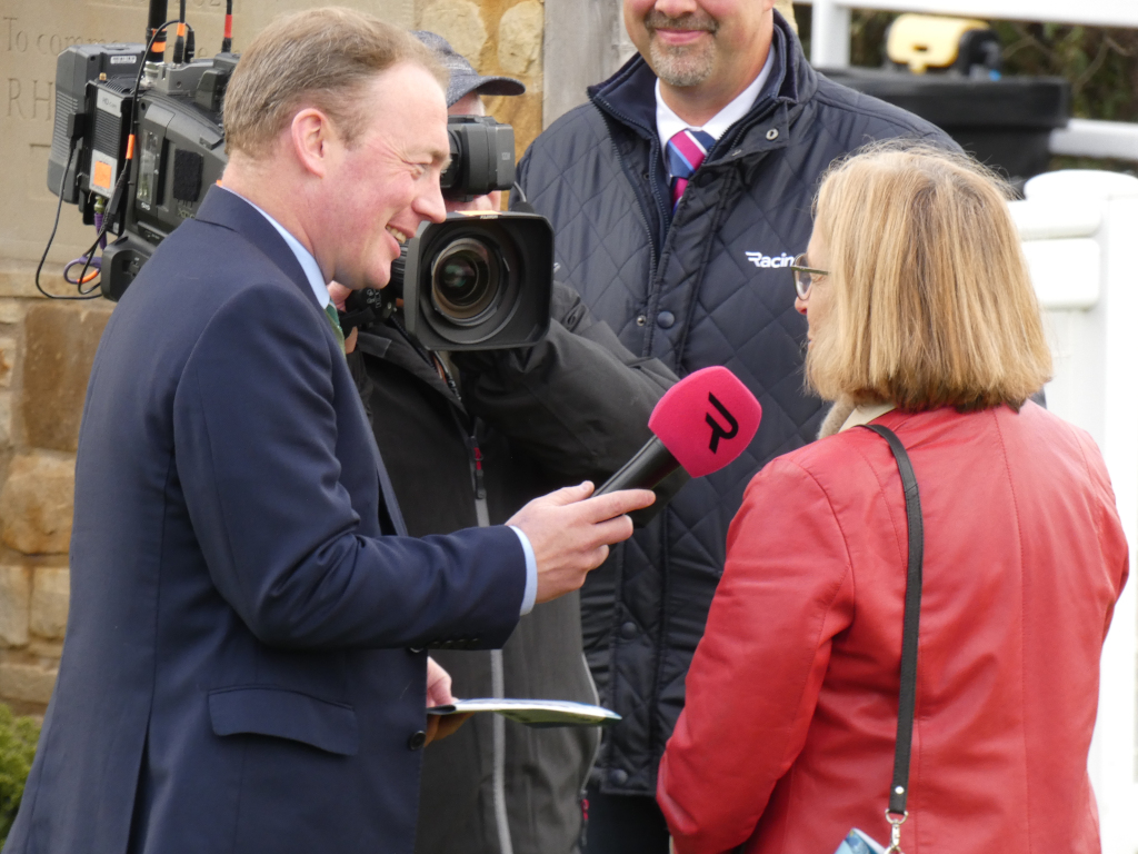 Man in a dark suit being interviewed by a woman wearing a red jacket. The interview is taking place outdoors, with a cameraman and another man in the background. The woman holds a pink microphone with a logo on it. The overall setting appears to be a public event, possibly at a racecourse or similar location, judging by the man in the background's jacket.