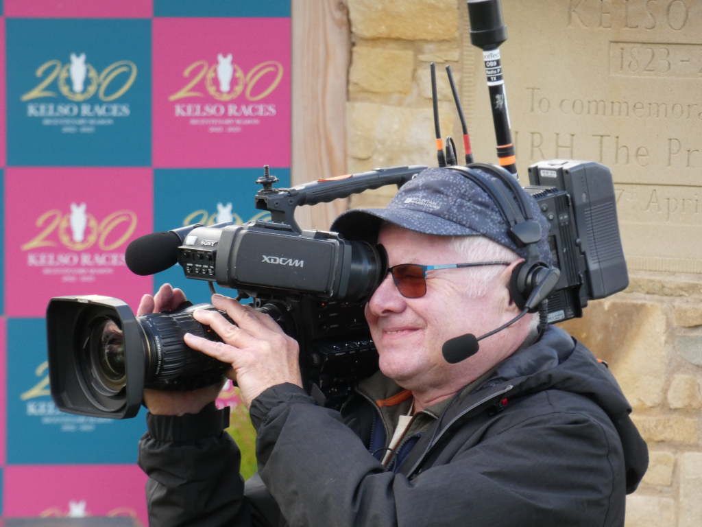 Cameraman, wearing a dark jacket, cap and sunglasses, operating a professional video camera. He is positioned in front of a backdrop displaying the logo for Kelso Races 200 which is repeated several times in a pink and teal grid pattern. In the background, a stone monument with an inscription is partially visible. The scene suggests the cameraman is filming an event at Kelso Racecourse, perhaps to commemorate its 200th anniversary. The image conveys a sense of professionalism and the behind-the-scenes work involved in documenting a significant event.