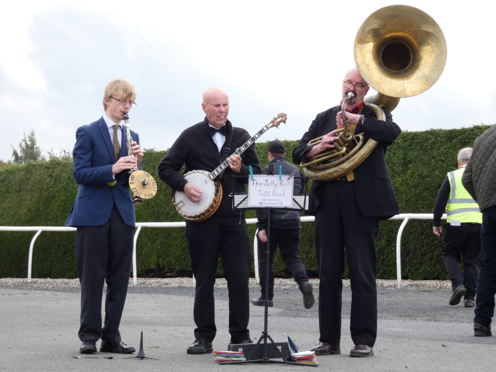 Three-piece jazz band performing outdoors, likely at a race track, judging from the fencing in the background. One musician plays a clarinet, another a banjo, and the third a baritone tuba or sousaphone. They are dressed in suits, giving a somewhat formal yet informal feel. A music stand with the band's name The Jelly Roll Jazz Band is positioned between the banjo and tuba players. Several onlookers are visible in the background, but they are out of focus. The overall impression is of a lively and impromptu performance in a slightly unexpected setting.