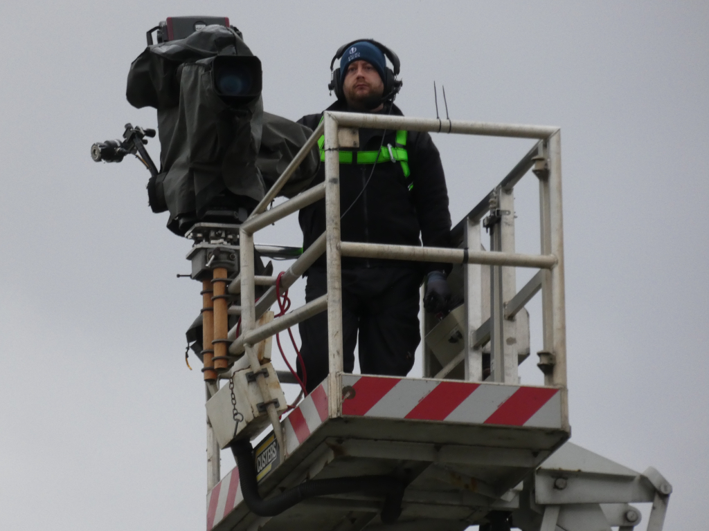 Cameraman in a lift, operating a large professional video camera. He's positioned high up against a grey sky, suggesting he's filming an outdoor event. The focus is on the cameraman and his equipment, conveying a sense of professionalism and the behind-the-scenes work involved in broadcasting or filming.