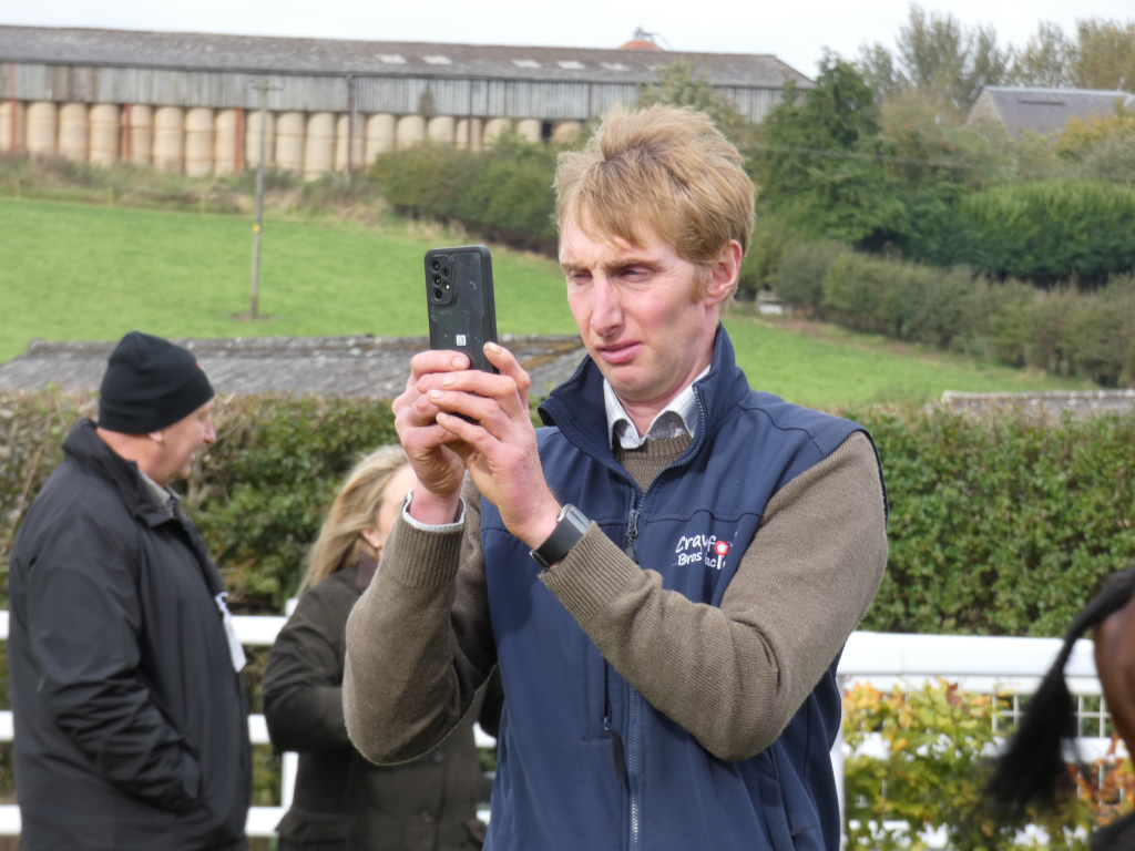Man with blonde hair, wearing a dark blue vest over a brown sweater, taking a picture with his smartphone. He appears to be in a rural setting, with farm buildings and fields visible in the background. Two other individuals are partially visible in the background, one wearing a dark jacket and the other a lighter-coloured jacket. The overall impression is of a casual, possibly professional, outdoor scene. The logo of Crave Brews is visible on his vest.