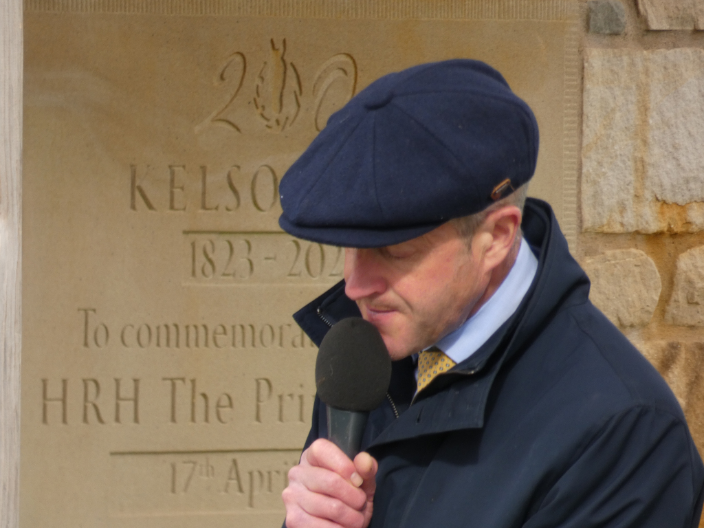 Man in a navy blue newsboy cap and dark jacket speaking into a microphone. He is standing in front of a light beige stone monument with an inscription commemorating HRH The Prince and the years 1823-2023. The inscription also includes the word Kelso and the date 17th April. The man seems to be giving a speech or commentating on the event. The overall mood is formal and commemorative.