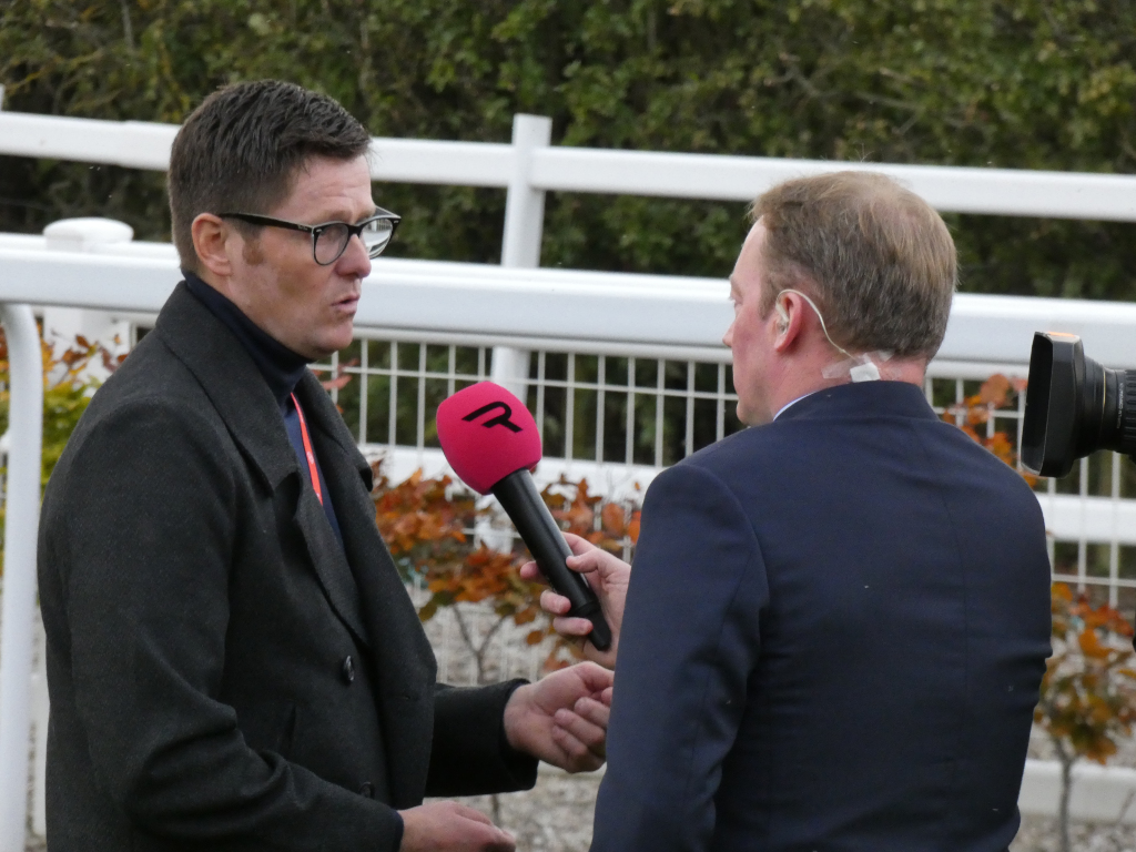 Man in a dark coat being interviewed by a reporter. The reporter holds a microphone with a pink R logo. The background features a white fence and some autumn foliage. The setting appears to be outdoors, possibly at a sporting event or similar public occasion.