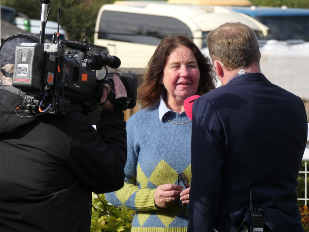 Woman being interviewed by a reporter. The woman is the main focus, wearing a blue and yellow sweater. A cameraman is visible in the background, recording the interview. The setting appears to be outdoors, near what looks like parked vehicles (possibly vans or buses). The overall impression is of a news interview or similar media event.