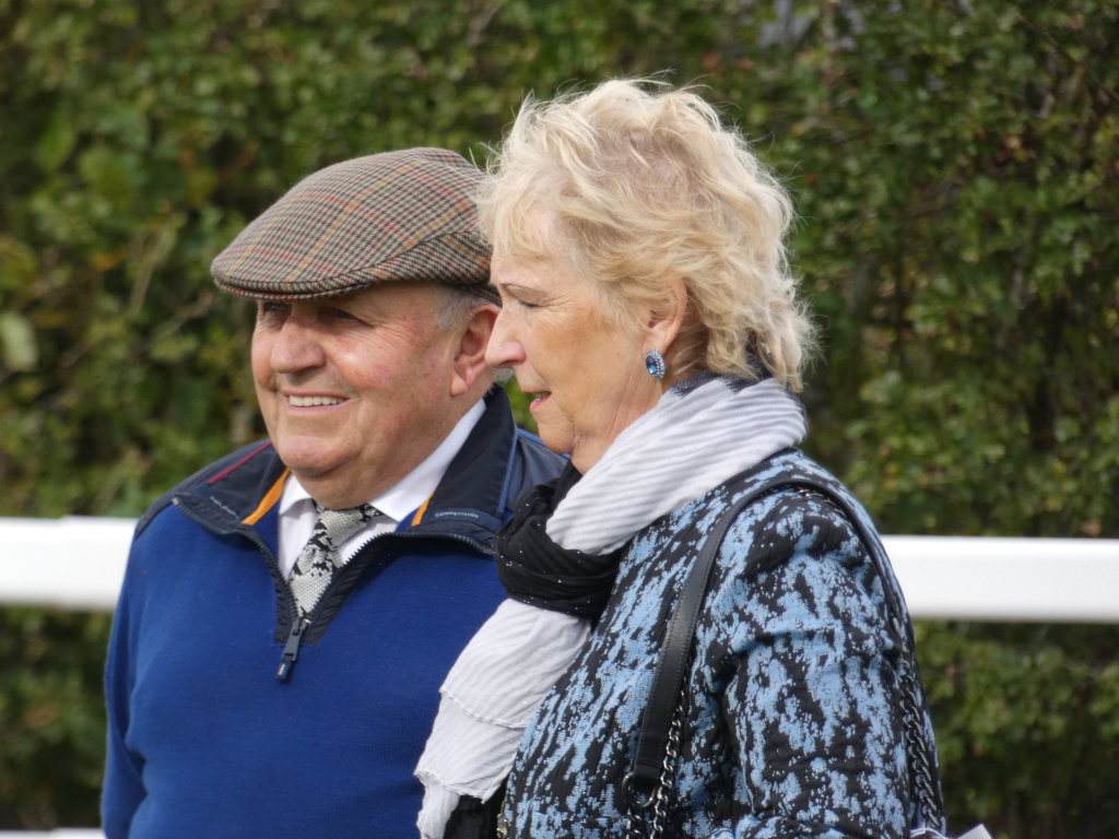 Close-up view of an older couple, positioned outdoors against a backdrop of lush green foliage. The man, wearing a tweed cap and a blue sweater, stands slightly behind and to the left of the woman. The woman, with short, blonde hair, is dressed in a patterned blue jacket and a grey scarf. They appear to be in conversation or simply standing close together, suggesting a close relationship, likely a married couple. The overall atmosphere is peaceful and intimate. The setting suggests an outdoor event, possibly at a racetrack or country club, given the man's attire.